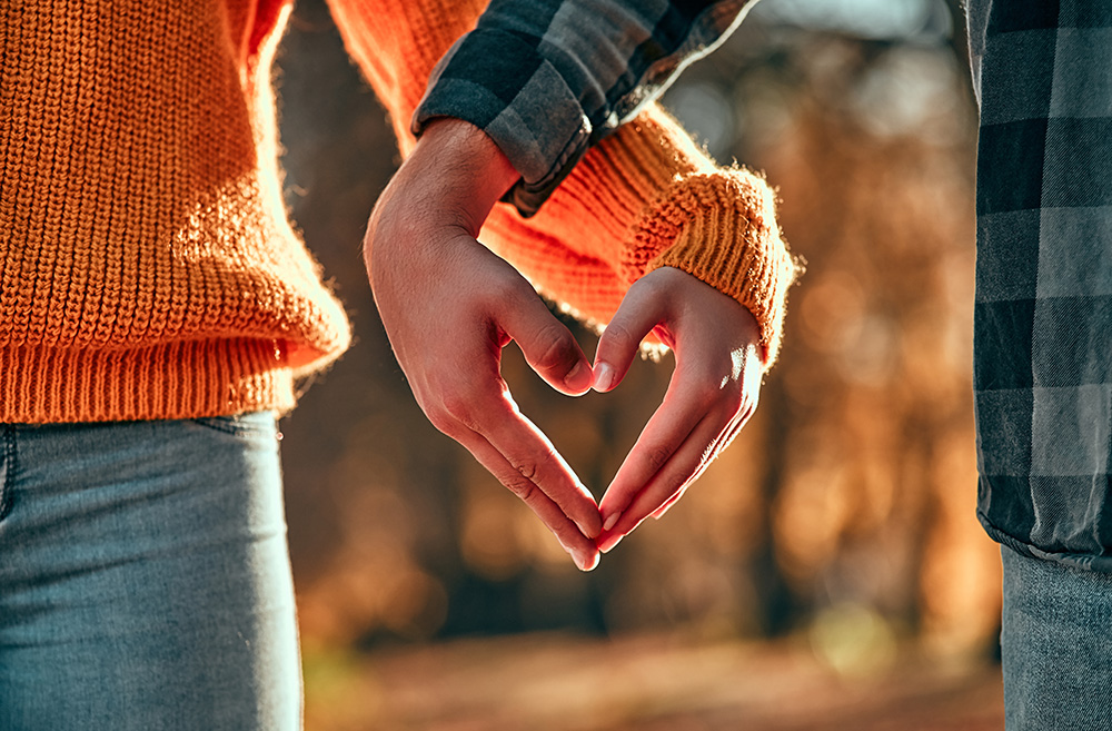 The hero image shows a close up of a man and a woman making the shape of a heart with their hands intertwined. Their lower arms, hands and parts of their torsos are visible only. The woman is wearing blue jeans and an orange sweater. The man is wearing blue jeans and a black and grey checked shirt.